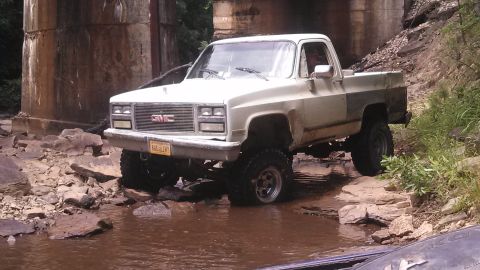 1979 Ford Bronco - A lifted, white GMC pickup truck is driving through a shallow, muddy creek bed near a concrete bridge structure. The truck has large off-ro…
