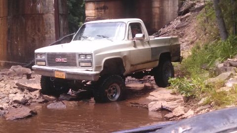 1979 Ford Bronco - A lifted, cream-colored GMC pickup truck with large off-road tires navigates a rocky stream bed under a concrete bridge. The license plate…