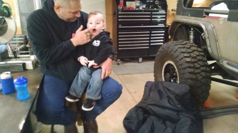1985 Jeep CJ7 - A man holds a baby in a garage setting, with a large off-road tire and part of a Jeep visible in the background. A toolbox and cans are als…