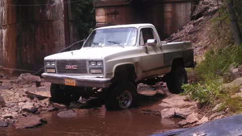 1979 Ford Bronco - A lifted, white GMC pickup truck is navigating a rocky, shallow stream under a concrete bridge. The truck has large off-road tires and a ye…