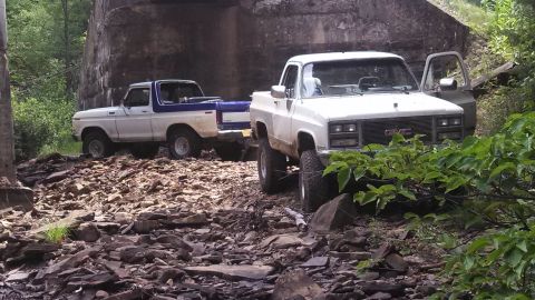 1979 Ford Bronco - Two off-road modified pickup trucks are parked on a rocky terrain next to a large concrete structure. The truck in the foreground is a whit…