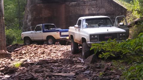 1979 Ford Bronco - Two vintage trucks are parked on a rocky terrain under a large concrete structure. A white and blue Ford Bronco is parked to the left, and…