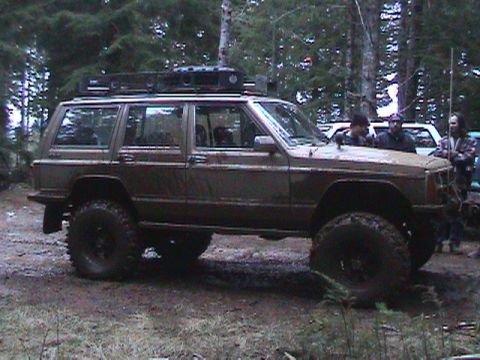 1994 Ford Explorer - Modified Jeep Cherokee XJ with large tires and roof rack parked in a forest.