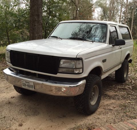 1996 Ford Bronco - A white 1996 Ford Bronco is parked on a dirt road surrounded by trees. The front of the SUV is facing the camera at a slight angle, showing…