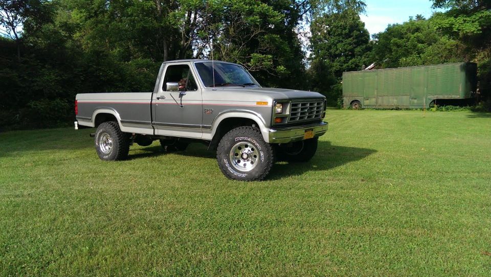 Lifted 1980s Ford F-250 pickup truck parked on grass with a green trailer in the background.