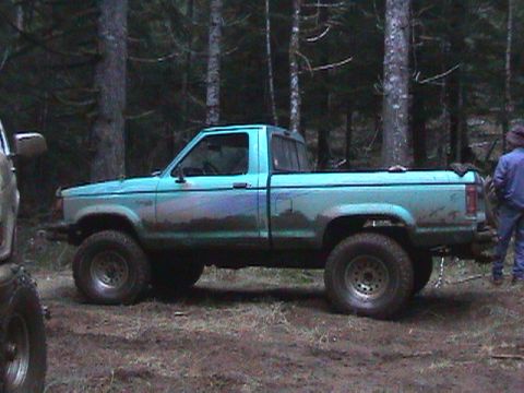 1994 Ford Explorer - A light blue pickup truck is parked off-road in a forest, with mud splattered on its sides.