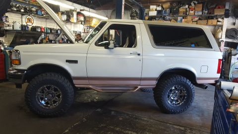 1995 Ford Bronco - Side view of a white Ford Bronco parked in a garage with the hood up. The vehicle has custom wheels, aftermarket side steps, and tinted win…