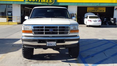 1995 Ford Bronco - Front view of a white 1990s Ford Bronco parked in front of a tire shop. The vehicle has a chrome grille, large off-road tires, and a Califo…