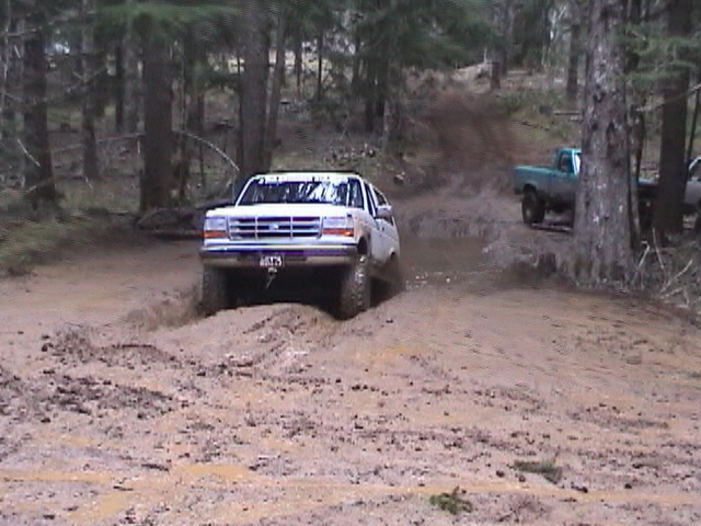 White Ford 4x4 driving through a muddy forest trail, teal pickup in the background
