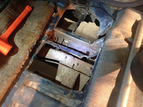 1979 Ford Bronco - The rusted-out floor pan of a vehicle is visible, with large holes revealing the undercarriage. An orange mallet lies on the carpeted floor…