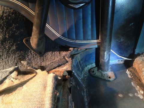 1979 Ford Bronco - A close-up view of the interior floor of a vehicle, showing rusted metal and carpet near the base of a roll bar support. The blue upholster…