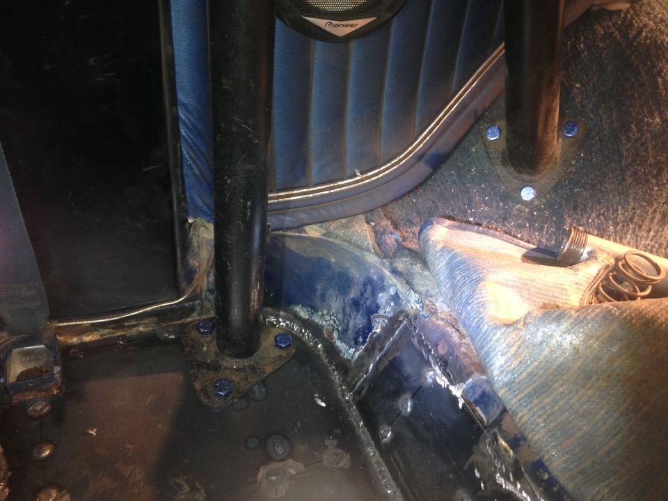 Vehicle interior floor with roll cage post, blue fasteners, and exposed metal.