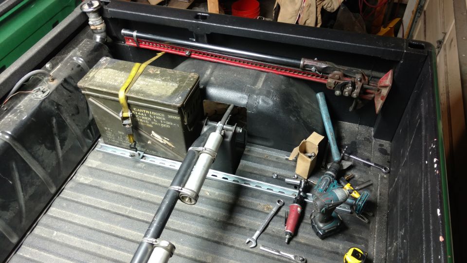 Pickup truck bed with tools including an ammo can, high-lift jack, drill, and fuel tank.