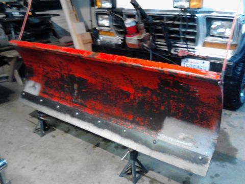1973 Ford F-250 - A red snow plow is mounted on the front of a truck. The plow is supported by jack stands and shows signs of wear and tear.