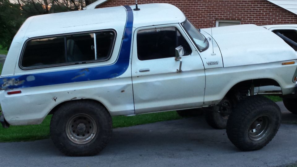 Lifted white 1979 Ford Bronco with blue stripe, side view, large off-road tires.