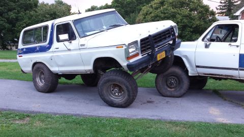 Two white Ford Broncos, one lifted and resting on the other, parked on pavement.