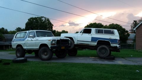 Two lifted SUVs, a Ford Bronco and a Chevrolet K5 Blazer, parked outdoors with a dramatic sunset sky.