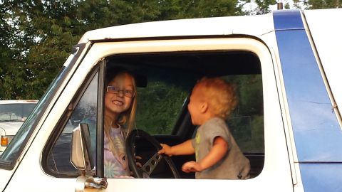 Two children in a white Ford Bronco, one in the driver's seat, the other reaching for the steering wheel.