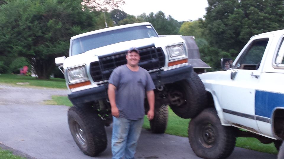Man standing between two lifted pickup trucks, one white, one white with blue stripe.