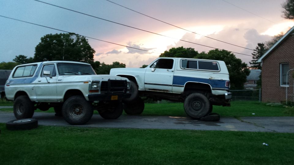 Two lifted SUVs, a Ford Bronco and a Chevy K5 Blazer, parked on a driveway with a tire on the grass.