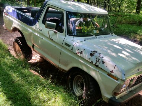 1979 Ford Bronco - A white 1979 Ford Bronco is shown from a low angle, driving off-road on a dirt path surrounded by lush green grass and trees. The vehicle h…