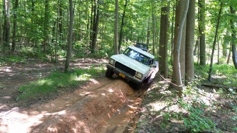 1979 Ford Bronco - A white 1970s Ford pickup truck is driving off-road on a muddy track through a forest. The truck is tilted to the right as it navigates a r…