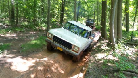 1979 Ford Bronco - A white 1970s Ford pickup truck is driving on a muddy trail through a forest. Another dark colored vehicle is visible behind it.