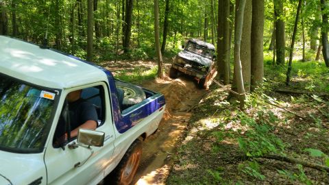 1979 Ford Bronco - Two off-road vehicles navigate a muddy trail through a dense forest. The foreground shows the rear and side of a white and blue pickup truc…