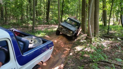 1979 Ford Bronco - Two off-road vehicles navigate a muddy trail through a dense forest. A dark-colored SUV is angled on the trail ahead, while the rear of a w…