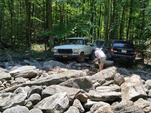 1979 Ford Bronco - A white 1970s Ford pickup truck navigates a rocky, wooded trail, with a black Jeep Grand Cherokee behind it. A person in shorts and a white…