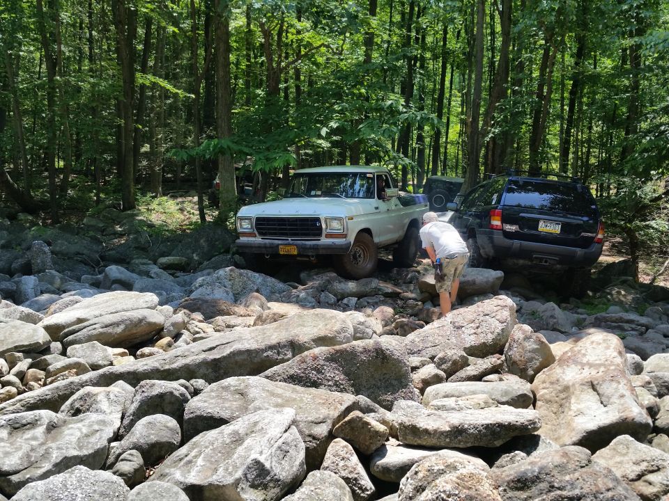 White 1970s Ford pickup truck and black Jeep Grand Cherokee on a rocky forest trail.
