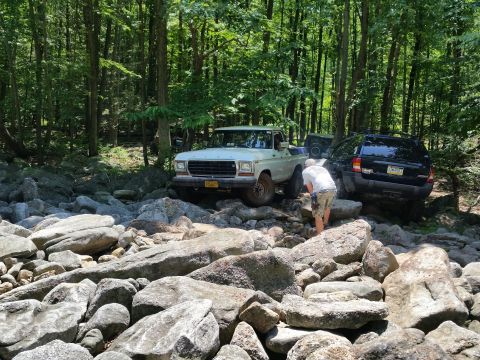 1979 Ford Bronco - A white 1970s Ford pickup truck is parked on a rocky, wooded trail next to a black Jeep SUV. A person in shorts and a t-shirt is standing b…