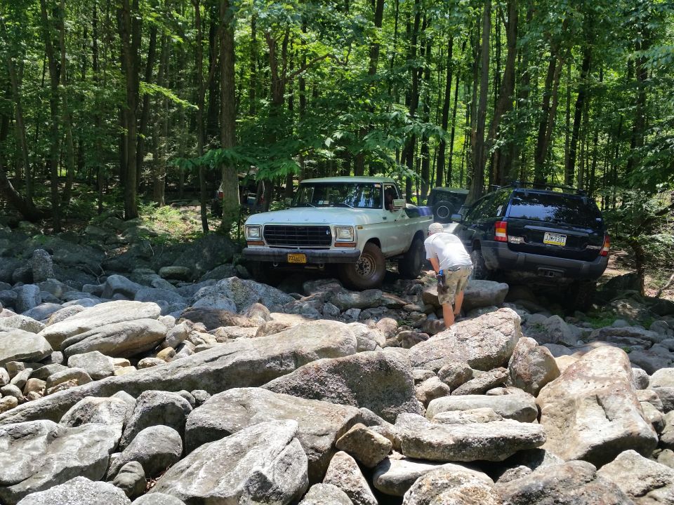 White 1970s Ford pickup truck and black Jeep SUV on a rocky forest trail with a person standing between them.