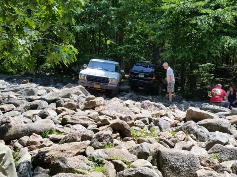 1979 Ford Bronco - A white 1979 Ford Bronco is parked on a rocky trail in a wooded area. A dark SUV is parked behind it, and two people are sitting on rocks t…