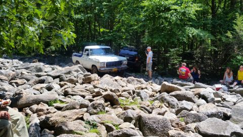 1979 Ford Bronco - A white and blue 1970s Ford pickup truck is parked on a rocky terrain next to a dark SUV. Several people are seated on rocks in the backgro…