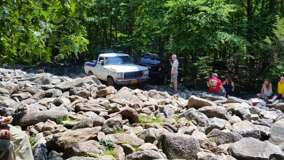 White and blue 1970s Ford pickup truck on rocky terrain with people in the background.