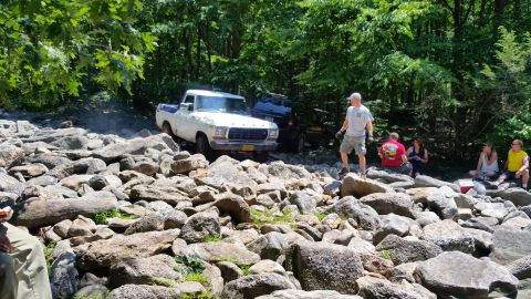 1979 Ford Bronco - A white and blue 1970s pickup truck is navigating a rocky terrain with a dark SUV behind it. Several people are gathered on the rocks watch…