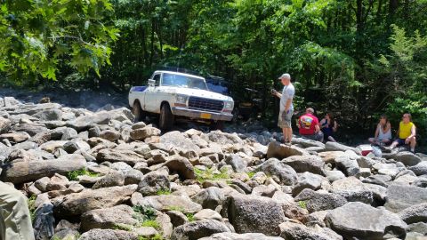 1979 Ford Bronco - A white 1970s Ford pickup truck is navigating a rocky terrain with a group of people watching in the background. The truck appears to be an…