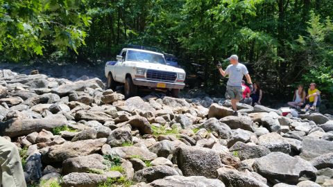 1979 Ford Bronco - A white and blue 1970s Ford pickup truck navigates a rocky, off-road terrain with trees in the background. Several people are gathered on t…