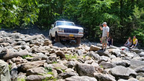 1979 Ford Bronco - A white 1970s Ford pickup truck is navigating a rocky, off-road trail with a group of people watching from the side. The truck appears to b…