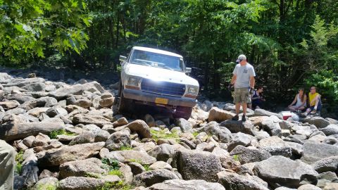 1979 Ford Bronco - A white 1970s Ford pickup truck navigates a rocky terrain with a group of people watching in the background. The truck is angled, with its…