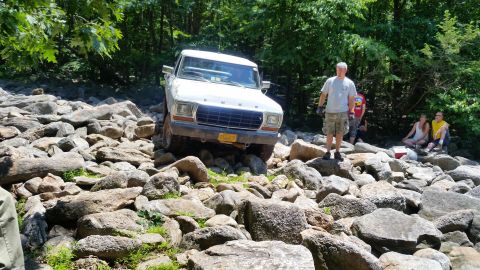 1979 Ford Bronco - A white 1970s Ford pickup truck navigates a rocky terrain with a group of people watching and resting nearby. The truck is positioned at an…