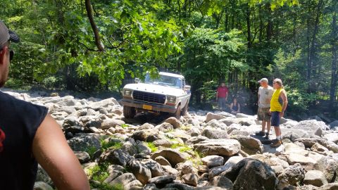 1979 Ford Bronco - A white 1970s Ford pickup truck navigates a rocky terrain in a wooded area. Several people are standing around watching the truck as it dri…
