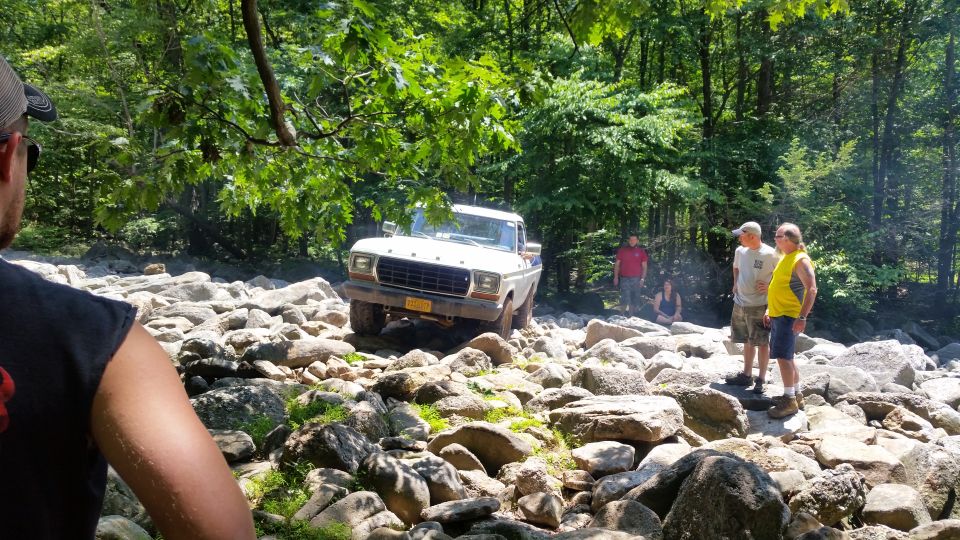White 1970s Ford pickup truck driving over rocks in a forest with people watching.