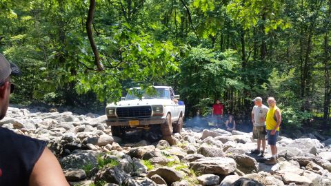 1979 Ford Bronco - A white 1970s Ford pickup truck navigates a rocky, wooded terrain. Several people are gathered around watching the off-road vehicle.