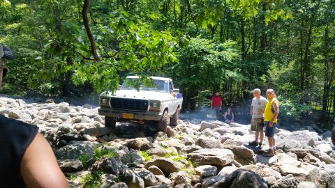 1979 Ford Bronco - A white 1970s Ford pickup truck is navigating a rocky, wooded terrain. Several people are standing around watching the off-road activity.