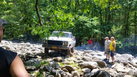 1979 Ford Bronco - A white 1970s Ford pickup truck is navigating a rocky terrain in a wooded area. Several people are standing around watching the vehicle.