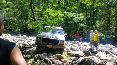 1979 Ford Bronco - A light-colored Ford truck navigates a rocky, wooded terrain with several people watching. The truck is positioned at an angle, with its fr…