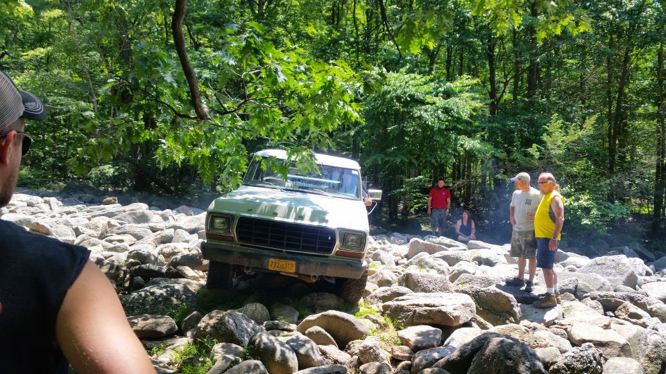 Light-colored Ford truck off-roading over large rocks in a wooded area with people observing.