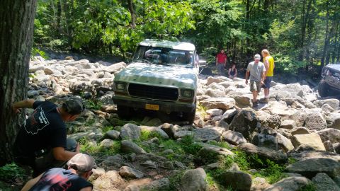 1979 Ford Bronco - A light-colored Ford Bronco navigates a rocky trail in a wooded area. Several people are visible around the vehicle, observing or assisting.